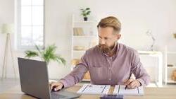 Middle aged blonde bearded man referring to laptop as he writes on papers in home office